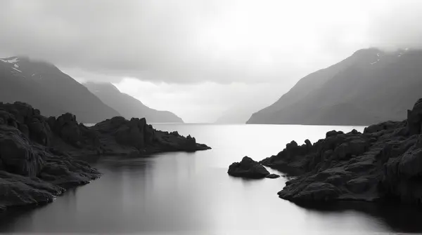 Black and white image of a smooth lake bordered by sharp stone formations under a cloudy sky