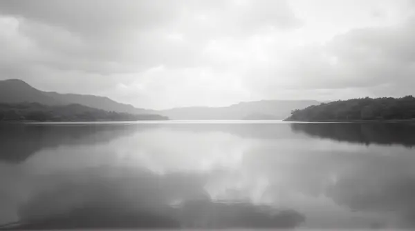 Black and white image of a lake reflecting a cloudy sky with tree silhouettes in the distance under soft light.