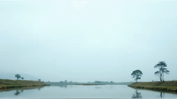 Calm lake bordered by green grass and scattered trees under a pale blue sky from a low angle