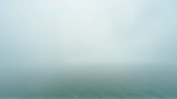 Vast blue-green sea stretching to horizon beneath soft white vapor clouds from a low angle