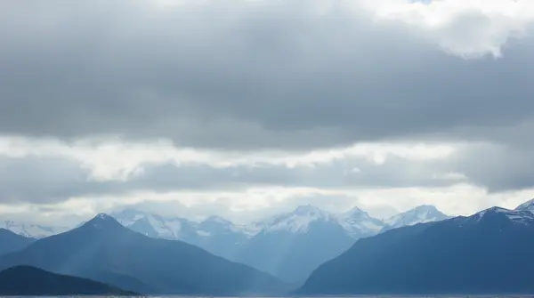 Wide view of towering peaks stretching across the horizon under textured gray clouds from a low angle