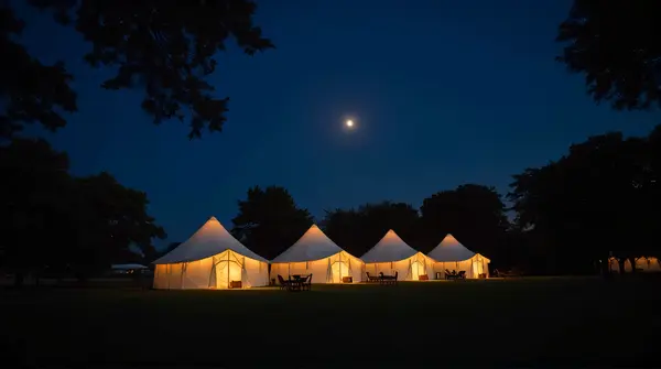 Cluster of softly lit tents on a grassy field with silhouetted trees and a moonlit sky