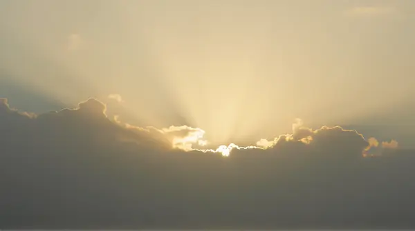 Golden rays shining partially hidden behind layered gray clouds creating soft shadows and muted tones across the wide sky.