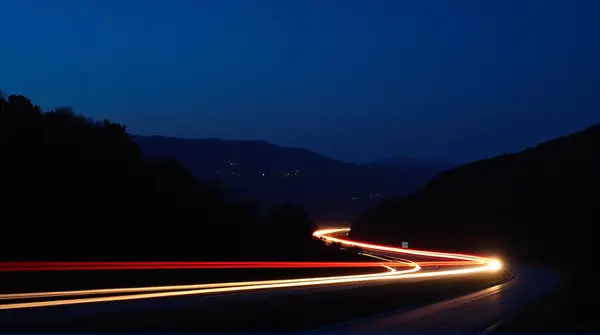 Long-exposure shot of a highway showing bright light trails from cars against dark hills and an indigo sky