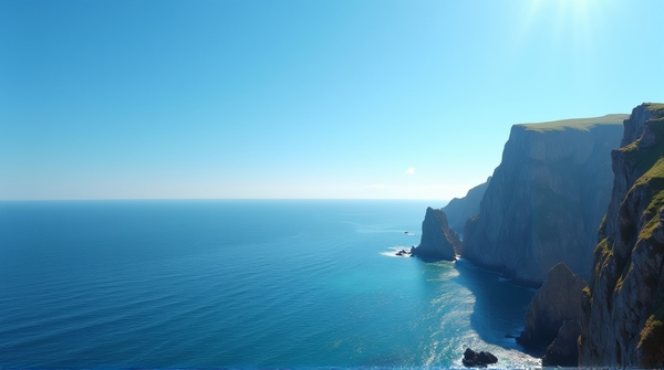 Jagged cliffs rise beside a calm ocean under a clear blue sky with sunlight casting sharp shadows near the coast.