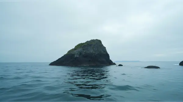 Massive stone pillar emerging from a wide sea with gentle ripples and cloudy sky above
