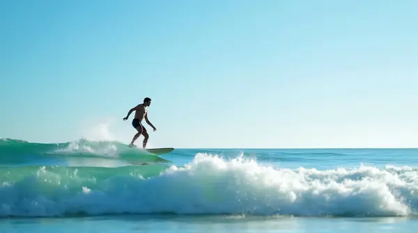 A man balancing on a surfboard glides over a cresting sea swell beneath a clear blue sky with distant horizon.