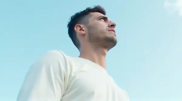 A man in a white long-sleeved top looking upward against a clear blue sky from a low-angle side view.