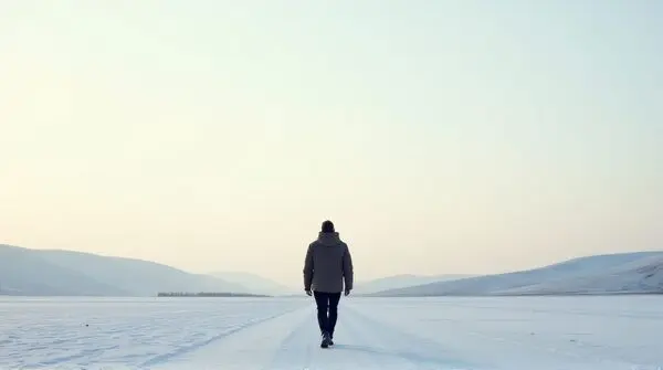 A man strides across a wide frost-covered plain under a pale sky with distant hills on the horizon.