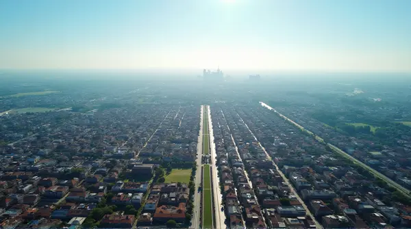 Bird's-eye perspective of a sprawling metropolitan area with rooftops, pathways, and green spaces under clear sunlight.