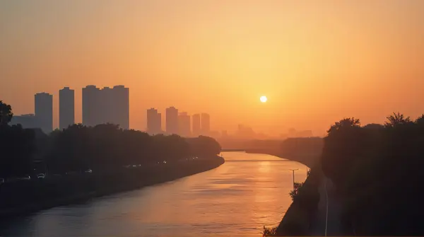 Silhouetted modern buildings against an amber horizon with a wide river in the foreground during soft fading light.