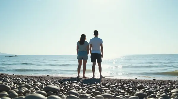 Two individuals stand on a pebble-lined shore beside gentle sea ripples under a vast sky at midday.