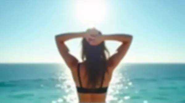 Person wearing a black bra stands with arms raised facing the sunlit ocean horizon under a clear blue sky.