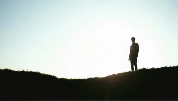 Person Standing On Grassy Rise Against Bright Sky