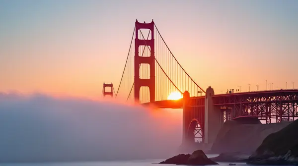 Low angle view of a red suspension bridge spanning a foggy bay at sunrise highlighting cables and towers.
