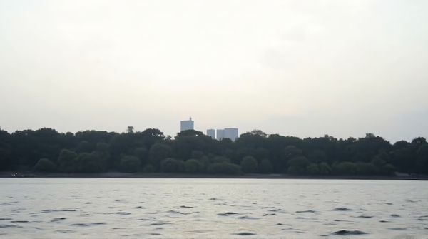 Dark outlines of buildings along a river with rippling water and soft clouds overhead in a pale sky