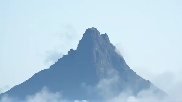 Low-angle view of a rocky summit partially covered by white mist under a pale blue sky.