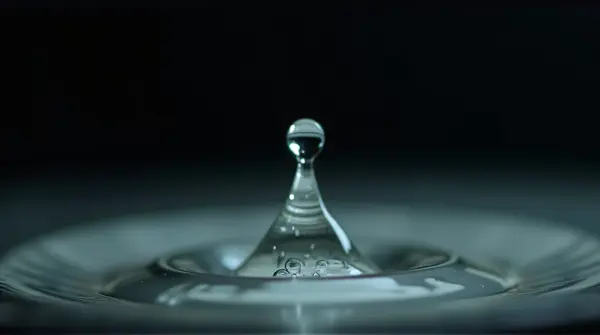 A sharp droplet hangs suspended midair reflecting bright surroundings against a dark background.