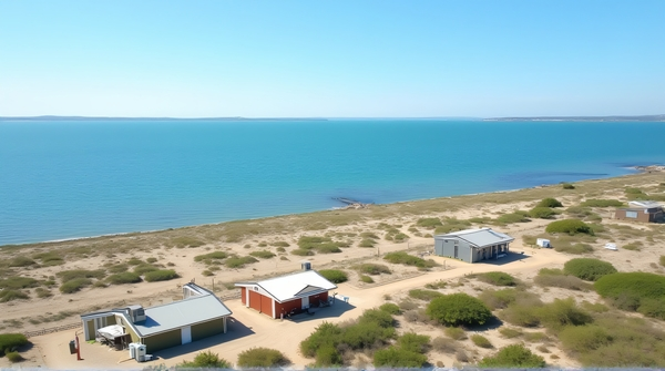 Bird’s-eye view of small buildings next to a large blue water body with sandy land and scattered plants under clear sky.