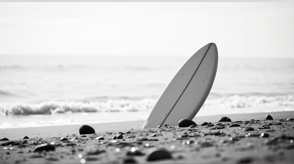 Monochrome image of a lone surfboard standing on a textured shoreline surrounded by seashells and gentle ripples in soft daylight.