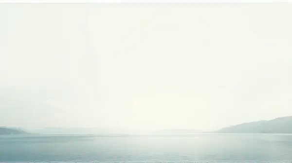 A wide still lake with soft light and distant low hills beneath a pale cloudless sky