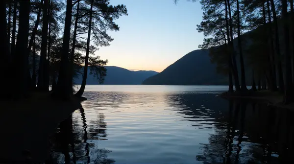 Dark outlines of tall trees beside a quiet lake with gentle ripples under a clear evening sky.