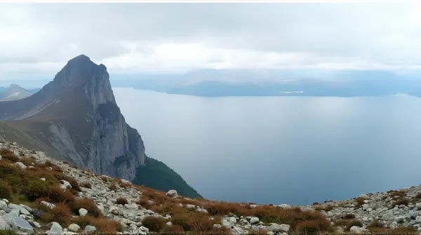 A towering peak rises next to a vast body of water under a cloudy sky with rocky terrain and alpine plants in the foreground.