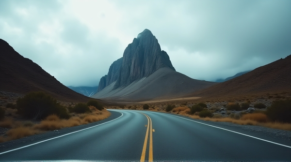 Low-angle view of a winding paved highway leading to a tall rocky peak under cloudy skies with sparse vegetation.