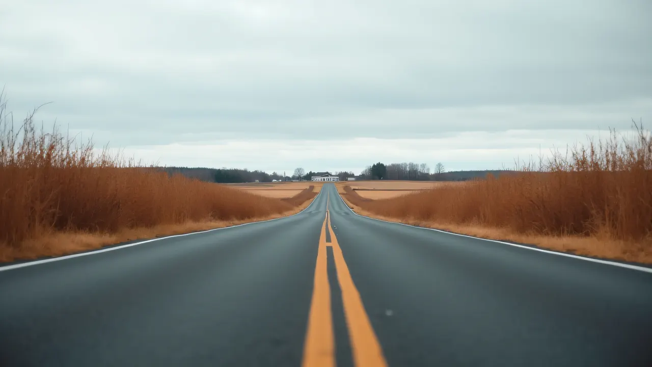Low-angle view of a straight asphalt road in a dry field with a farmhouse and cloudy sky.