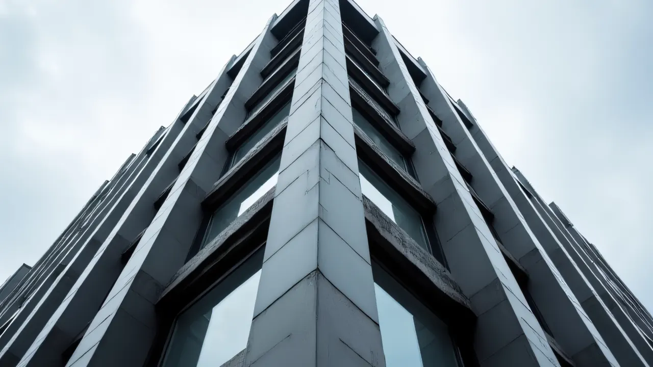 Low-angle view of a gray city building with strong lines and reflective windows under a cloudy sky.