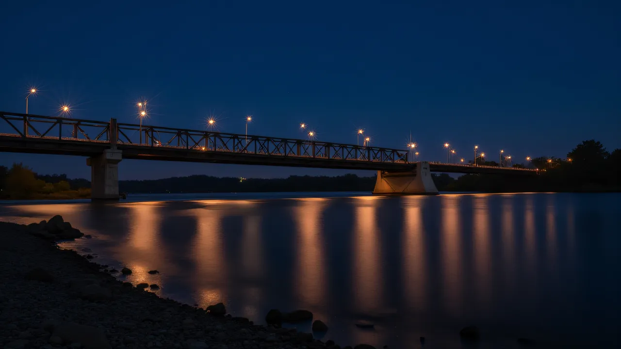 A low-angle view of a lit bridge reflecting in the calm river at night.