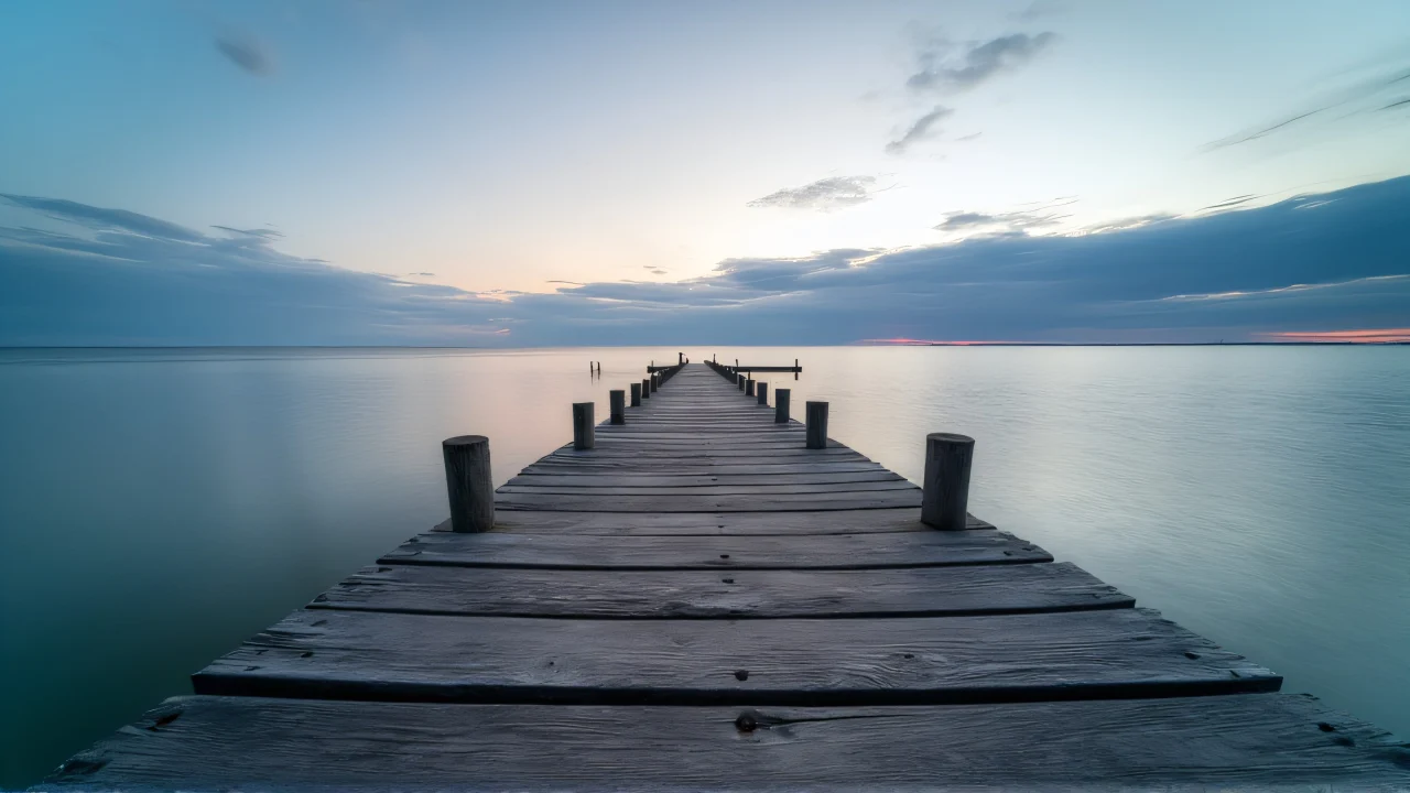 A weathered wooden pier stretches into calm water at dusk, leading to a soft horizon.